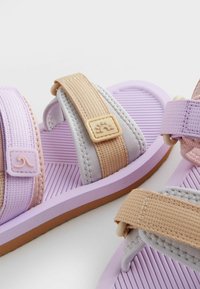 Violetinės ir smėlio spalvos šlepetės su elastingomis dirželiais, tekstūruotais padais ir dekoratyviais akcentais. Pagaminta iš kaučiuko ir audinio medžiagų.