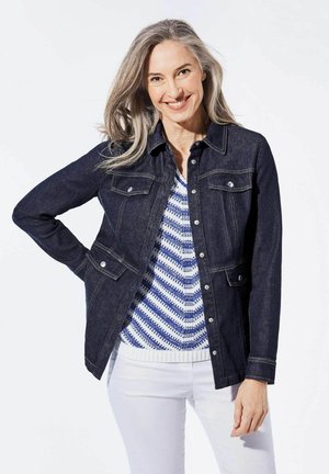 Veste en jean foncé avec un devant boutonné, deux poches poitrine et des coutures contrastées ; superposée à un pull à motifs bleu et blanc.