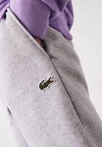 Pantaloni de trening gri din amestec de bumbac, cu un mic logo brodat al unui crocodil verde pe lateral. Materialul este moale și are o textură netedă.
