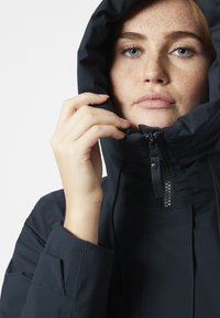 Černá mikina s kapucí, předním zipem, nastavitelnou šňůrkou a texturovanou tkaninou. Záběr na obličej osoby, která drží kapuci.