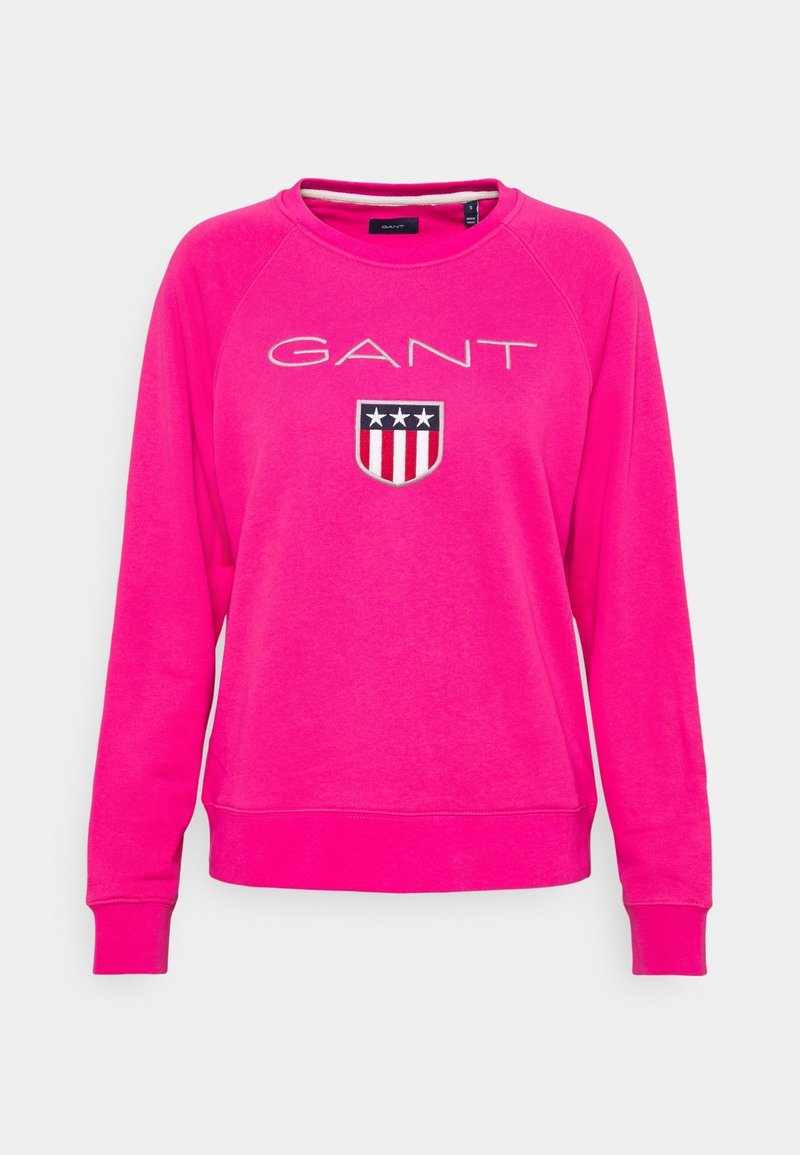 Sweat-shirt rose avec un col rond, des manches raglan, des poignets et un ourlet côtelés. Présente le logo "GANT" et un écusson brodé avec des étoiles et des rayures.