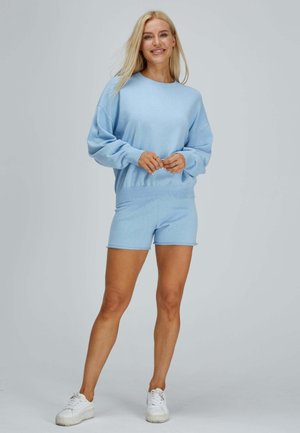 Lächelnde Frau stehend, trägt hellblauen Langarmtop und passende Shorts mit weißen Sneakers vor einfarbig grauem Hintergrund.