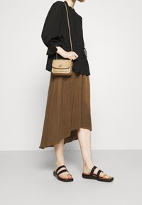Blouse noire texturée avec des manches froncées, jupe asymétrique marron et sandales noires à deux brides. Sac bandoulière beige avec quincaillerie dorée.