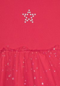 Crvena haljina s rebrastim gornjim dijelom, blistavim dizajnom zvijezda od rhinestones, i prozirnim volanima u donjem dijelu ukrašenim šarenim šljokicama u obliku zvijezda.