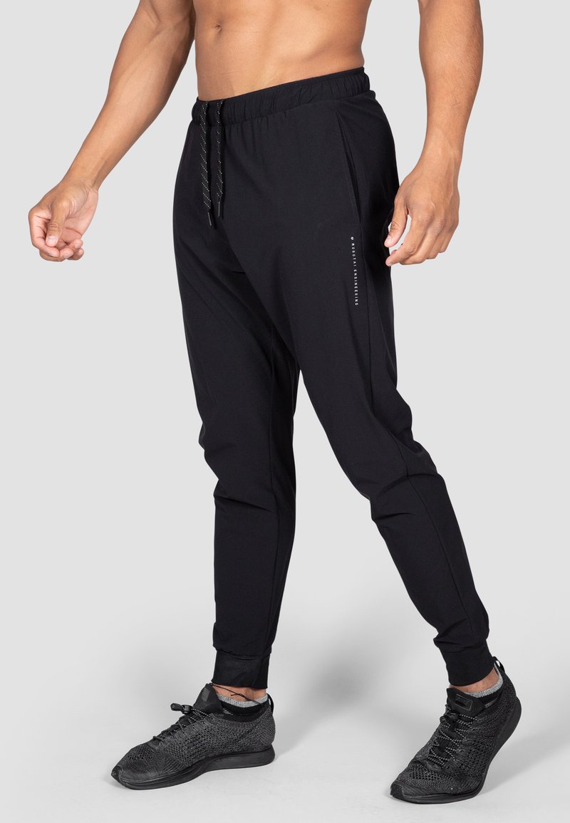 Schwarze Fitness-Jogginghosen mit elastischem Bund, Kordelzug und gerippten Bündchen. Mit Reißverschlusstaschen und einem reflektierenden Logo an der Seite.