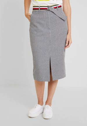 Mujer vistiendo una falda lápiz de pata de gallo en blanco y negro con abertura central, cinturón a rayas, zapatillas blancas y camisa blanca metida por dentro.