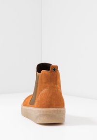 Spalone pomarańczowe zamszowe botki za kostkę z elastycznym panelem bocznym, przeszyciami, pętlą z oczkiem oraz teksturowaną jasną gumową podeszwą, ustawione w białym rogu.