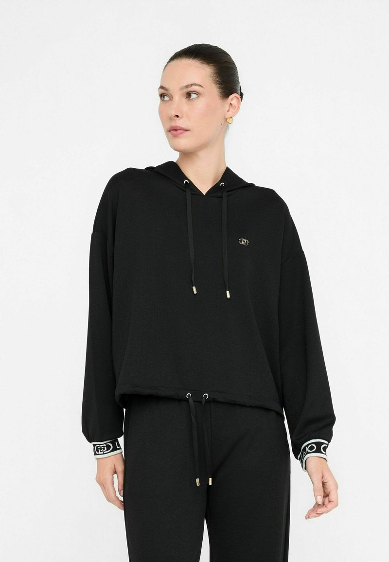 Schwarze, verkürzte Kapuzenjacke aus weichem Stoff, mit Kapuze, goldfarbenen Kordeln und gemusterten Bündchen. Einfaches Design mit gesticktem Logo.