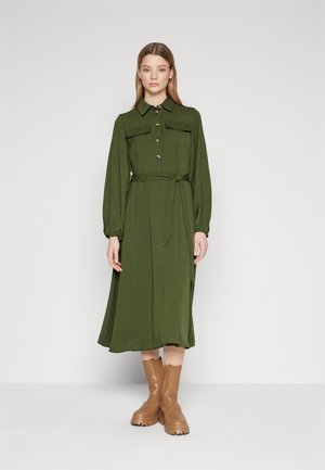 Long-sleeved, olive green dress with two chest pockets, button closure, and a tied waist belt. Paired with tan knee-high boots.