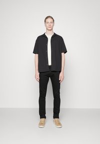 Schwarzes kurzärmliges Hemd über einem weißen T-Shirt, kombiniert mit schwarzen Skinny Jeans und beigen Sneakers; minimalistisches Design und neutrale Farben.