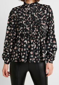 Blusa nera floreale con pieghe, caratterizzata da piccoli fiori rosa e verdi. Maniche lunghe con polsini elasticizzati, design a collo alto. Tessuto dalla texture liscia.