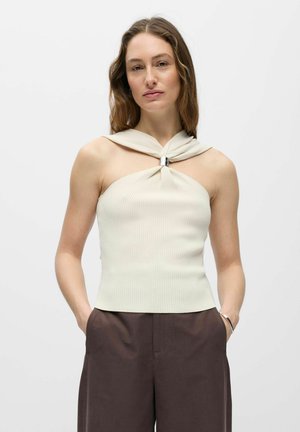 Hosszú hajú nő, aki ujjatlan bézs felsőt visel csavart nyakkivágással és sötétbarna nadrággal, kezeit zsebre téve áll.