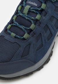 Scarpa da trekking blu navy con materiali in rete e suede, lacci verdi riflettenti e suola grigia testurizzata con motivi di battistrada profondi.