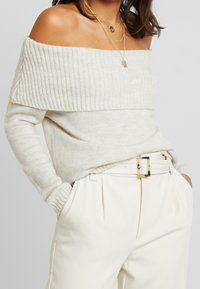 Cremfarbener schulterfreier Pullover aus weichem Strickmaterial mit gerippter Textur am Halsausschnitt, kombiniert mit hochtaillierten cremfarbenen Hosen und einem Gürtel.