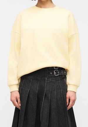 Personne portant un pull jaune pâle à manches longues rentré dans une jupe noire plissée taille haute avec des détails à double boucle.