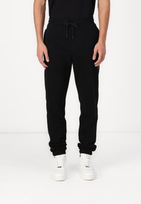Schwarze Jogginghose mit einem Kordelzugbund, schmal zulaufenden Beinen und seitlichen Taschen. Kombiniert mit weißen Sneakers für einen Kontrast.