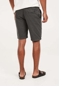 Shorts en coton gris avec une coupe droite, poche arrière avec rabat, portés avec des tongs noires. Design simple et décontracté.