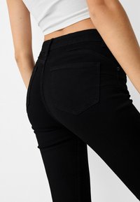 Mujer vistiendo jeans skinny negros de cintura alta y una blusa corta blanca, vista desde atrás contra un fondo sencillo.