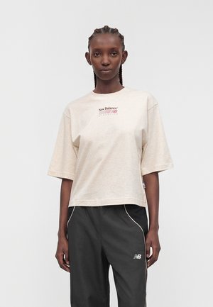 Mujer con el pelo trenzado que lleva una camiseta beige de New Balance Athletics y pantalones de chándal negros de New Balance, de pie frente a un fondo liso.