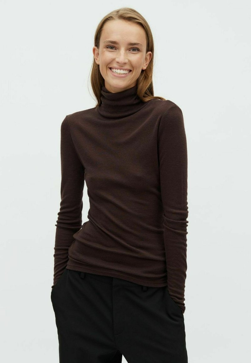 Femme souriante aux longs cheveux, portant un col roulé marron foncé et un pantalon noir, debout les mains dans les poches devant un fond clair uni.