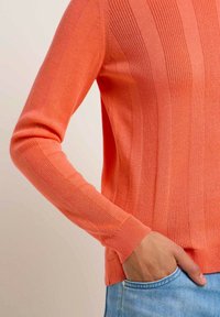 Ribbed orange sweater with long sleeves and fitted cuffs, made of textured knit material; shown with a hand in a blue denim pocket.
