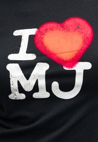 Μαύρο πουκάμισο με λευκό κείμενο "I" και "MJ" χωρισμένα από μια κόκκινη καρδιά με ένα μικρό μαύρο λογότυπο Jumpman στην πάνω δεξιά γωνία της καρδιάς.