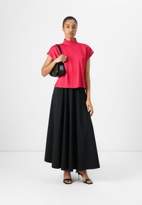 Fitted bright pink top with mock neck and short sleeves, paired with a long black pleated skirt. Model holds a small black handbag.