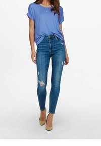 Fioletowy T-shirt z krótkim rękawem, wysokiej talii niebieskie dżinsy z efektami postarzania oraz cieliste buty na wysokim obcasie. Dżinsy mają przetarcia na kolanach.