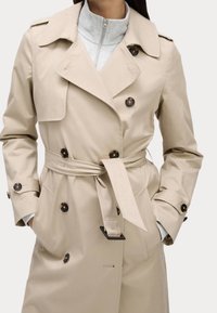 Beżowy płaszcz trencz o dwurzędowym kroju, z dużymi brązowymi guzikami, paskiem i naramiennikami. Materiał wydaje się miękki i gładki.