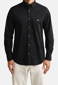 Homme portant une chemise noire à manches longues boutonnée avec une poche sur la poitrine et un pantalon beige, debout avec les bras détendus le long du corps.