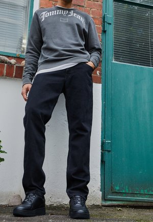 Person steht im Freien und trägt einen grauen Tommy Jeans Pullover, schwarze Hosen und schwarze Turnschuhe neben einer grünen Tür und einer Backsteinwand.