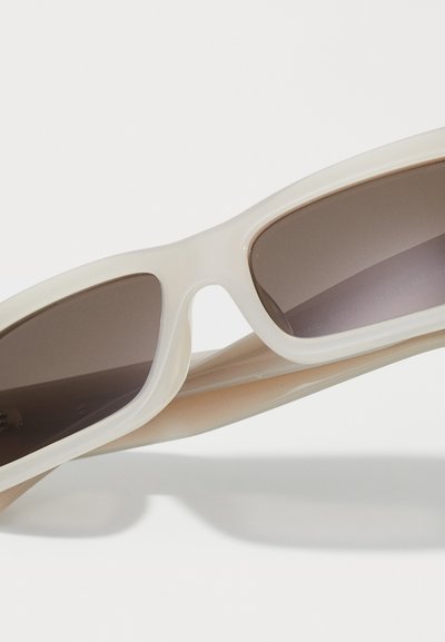 Lunettes de soleil rectangulaires couleur crème avec des verres teintés foncés reposant sur une surface blanche.