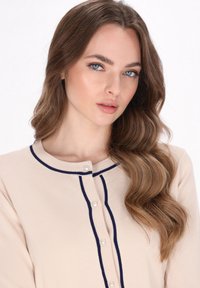 Beige cardigan met een ronde halslijn, marineblauwe afwerking en pareldoppen. Het materiaal heeft een textuurafwerking en de mouwen zijn iets losjes passend.