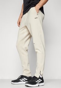 Beige sweatpants with side pockets, elastic waistband, and logo detail. Worn with black sneakers featuring white stripes.