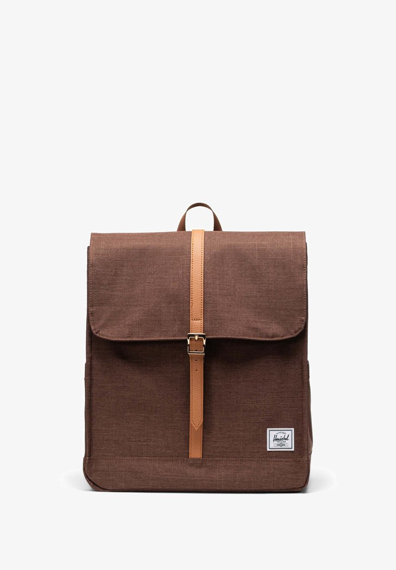 Brauner Stoffrucksack mit Deckelklappe, hellbraunem Ledertragegurt und Schnallenverschluss sowie einem Logo-Patch auf der Vorderseite. Rechteckige Form, strukturiert Oberfläche.