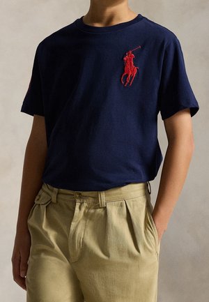 Person iført navyblå kortærmet skjorte med rød polospillerlogo, kombineret med beige plisserede bukser og hænderne i lommerne.