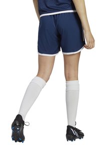 Marineblaue Sportshorts mit weißem Saum, kombiniert mit weißen kniehohen Socken und schwarzen Stollen mit blauen Akzenten, von hinten betrachtet.