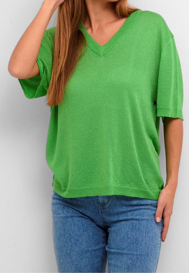 Femme portant un haut vert vif à manches courtes et col en V, et un jean bleu, une main touchant ses cheveux et l'autre reposant sur le côté.