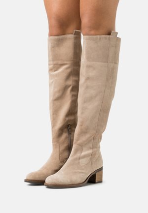 Tan suède kniehoge laarzen met blokhakken en zijritsen, gedragen op benen tegen een witte achtergrond.