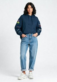 Marineblauwe hoodie met een koord, voorzien van kleurrijke geborduurde ontwerpen op de mouwen. Gecombineerd met lichtblauwe, opgerolde jeans en witte sneakers.
