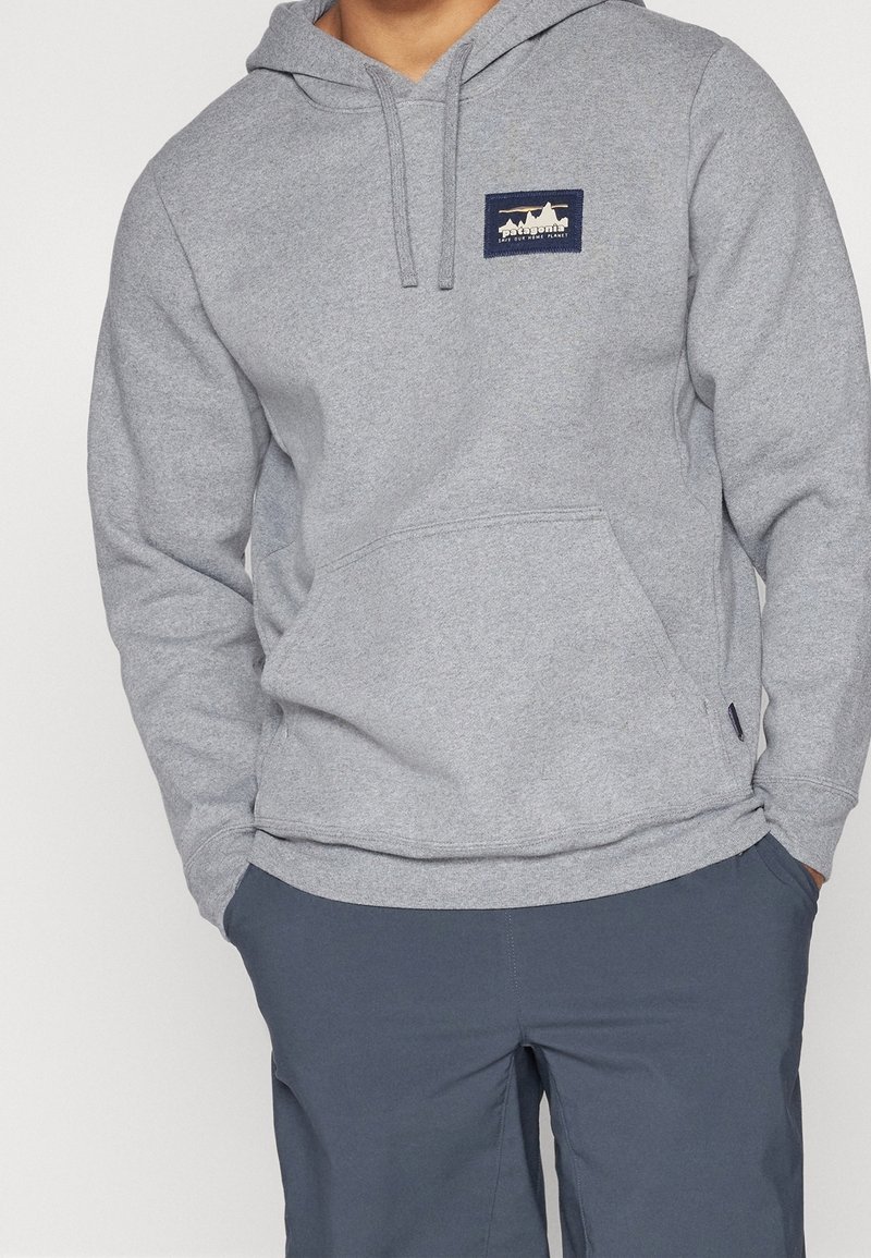 Personne portant un sweat à capuche Patagonia gris clair avec une poche avant et un pantalon bleu marine, mains dans les poches.