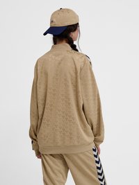 Ženska v svetlo rjavi trenirki s vzorcem in ujemajoči se bež in mornarsko modri kapici, stoji s hrbtom proti kameri na enobarvnem ozadju.