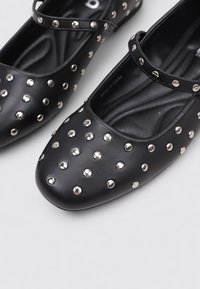 Bailarinas de cuero negro con punta redonda, con remaches metálicos dispuestos decorativamente en la superficie superior y una correa cruzando el empeine.