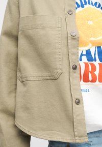 Μπεζ σακάκι με κουμπιά και μπροστινή τσέπη πάνω από λευκό T-shirt με φέτες πορτοκαλιού και μπλε και κόκκινο κείμενο.