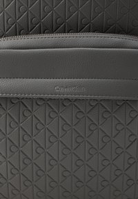 Grå læderhåndtaske med et struktureret geometrisk mønster, et glat horisontalt panel og præget logo detalje. Lynlåslukning på toppen.