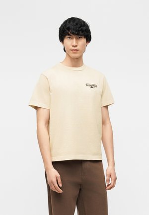 Fiatal férfi fekete hajjal, bézs pólót visel, amelyen a "Service Works" felirat olvasható a mellkasán és barna nadrágot, egy egyszínű háttér előtt áll.