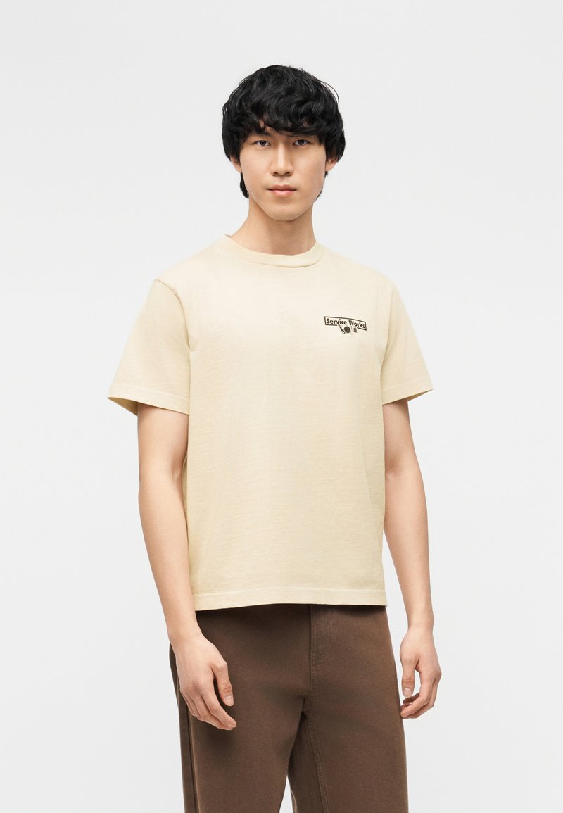 Νέος άνδρας με μαύρα μαλλιά που φοράει μπεζ μπλουζάκι με το κείμενο «Service Works» στο στήθος και καφέ παντελόνι, στέκεται μπροστά σε απλό φόντο.