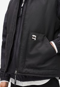 Melna izolēta veste ar rāvējslēdzējslēgumu, kas aprīkota ar kabatu ar uzplecu un dekoratīvu nozīmi. Materiāls izskatās gluds ar elastīgiem manžetēm.