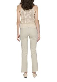 Débardeur en crochet beige avec un motif ouvert et des bretelles fines, associé à un pantalon ajusté beige clair. Le modèle est de dos, mettant en valeur la tenue.