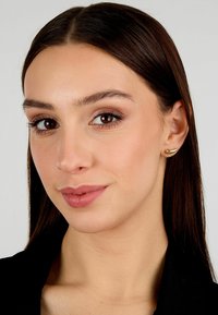 Orecchini a vite dorati con un design curvo, texture liscia e finitura lucida, indossati da una persona con lunghi capelli scuri e trucco neutro.